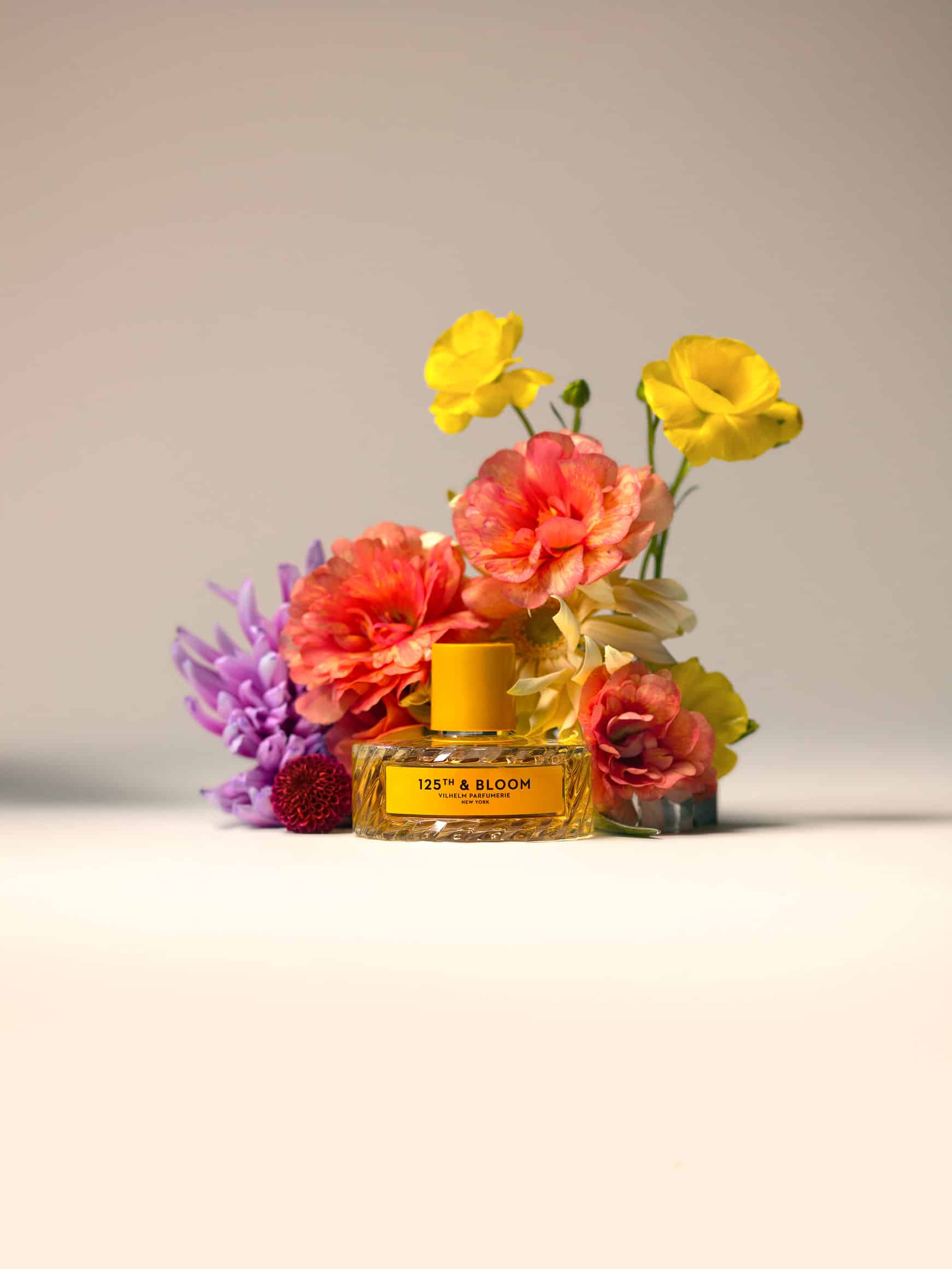 Vilhelm Parfumerie 12th and Bloom perfume bottle styled with yellow, pink, purple, and peach flowers on a white background