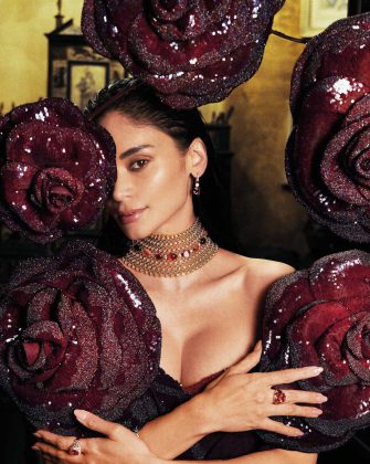 Pia Wurtzbach-Jauncey posing behind large, sculptural deep red roses while wearing a gold Bulgari choker with red gemstones.