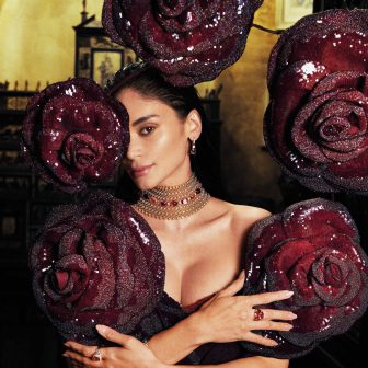 Pia Wurtzbach-Jauncey posing behind large, sculptural deep red roses while wearing a gold Bulgari choker with red gemstones.