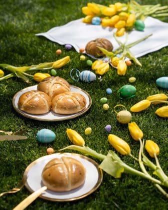 A spring Easter picnic setup on green grass featuring freshly baked hot cross buns on gold-rimmed plates, yellow tulips, and colorful Easter eggs for the Sorbet 2026 Guide.