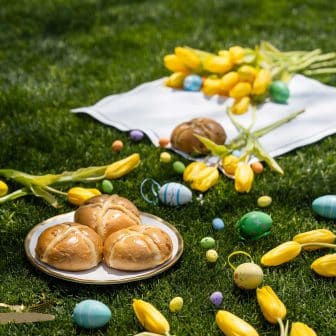 A spring Easter picnic setup on green grass featuring freshly baked hot cross buns on gold-rimmed plates, yellow tulips, and colorful Easter eggs for the Sorbet 2026 Guide.