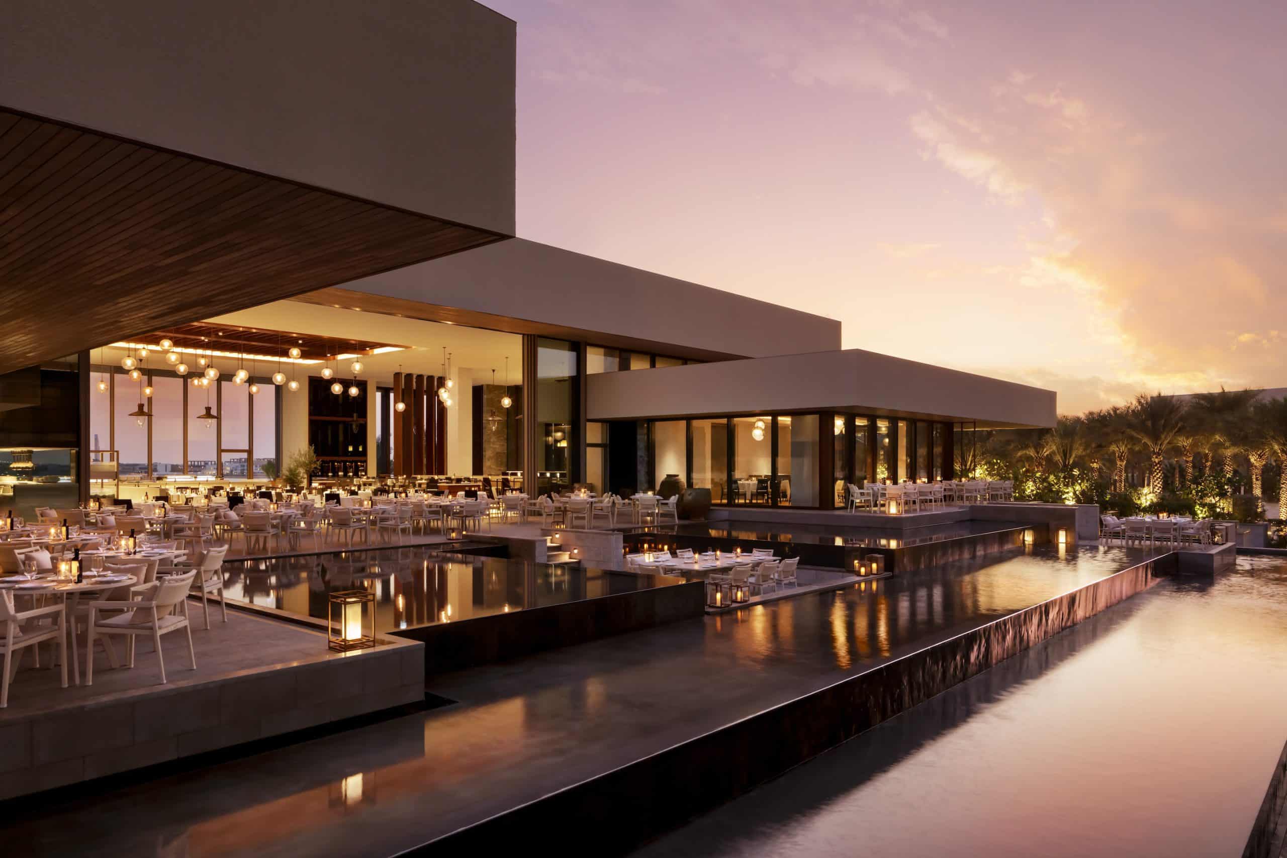 Twilight view of the ultra-luxury estiatorio milos Atlantis The Royal resort in Dubai, showing the modern stacked-block architecture, glowing restaurant interiors, and infinity pools reflecting a purple sunset.