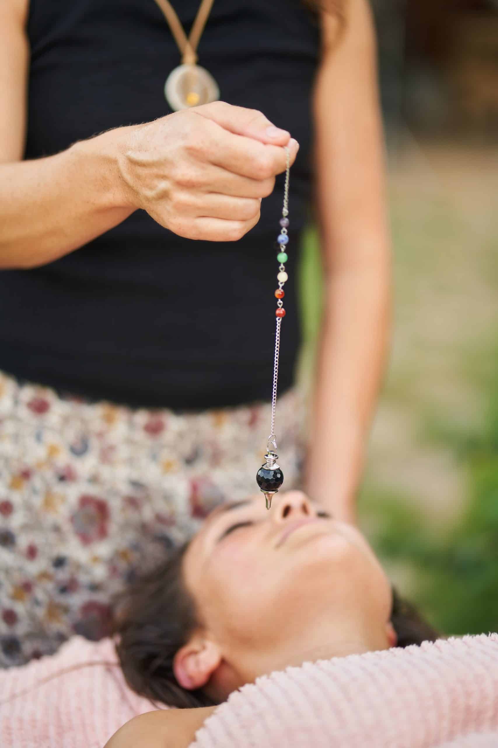 Therapist holding pendulum over young woman during crystal healing therapy session promoting relaxation and spiritual balance